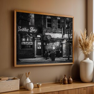 May include: Black and white framed photograph of a building with the neon sign "Cadillac Lounge". The image shows a vintage car mounted on the building's facade. The photograph is displayed on a brown wall above a wooden cabinet.