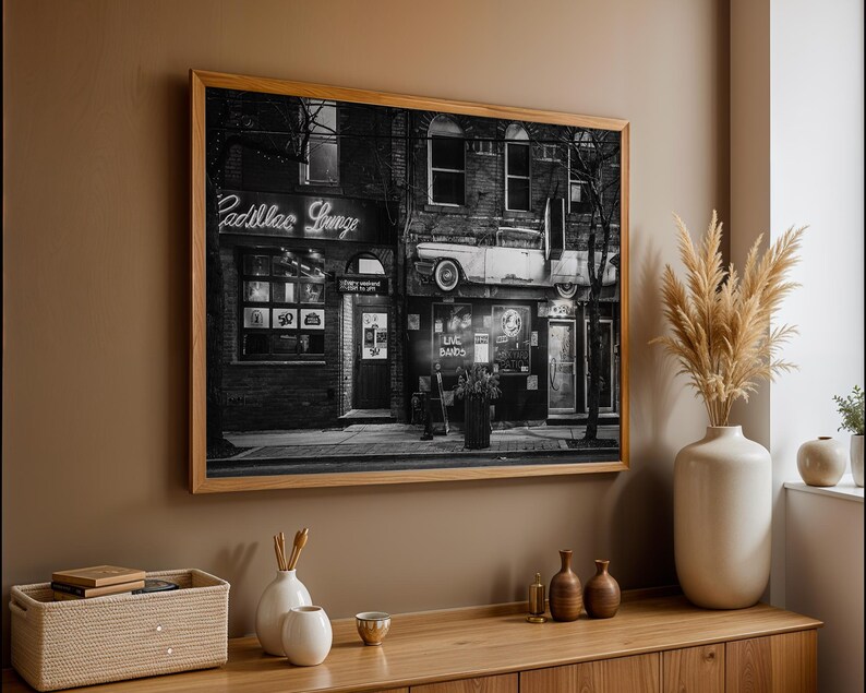 May include: Black and white framed photograph of a building with the neon sign "Cadillac Lounge". The image shows a vintage car mounted on the building's facade. The photograph is displayed on a brown wall above a wooden cabinet.
