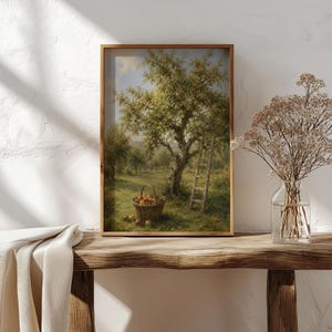 May include: A framed wall art print depicting an orchard scene with a large apple tree, a wooden ladder leaning against the trunk, and a woven basket filled with red apples on the grass. The artwork features soft green, brown, and red tones.