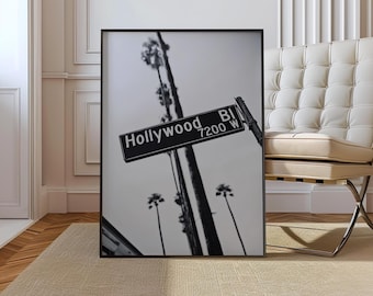 May include: Black and white framed print featuring a Hollywood street sign. The sign reads "Hollywood BI 7200 W" and is set against a backdrop of palm trees and a cloudy sky. The print is displayed on a beige rug.