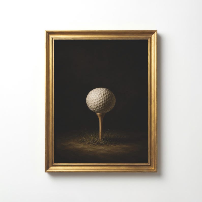 Op de afbeelding: Een ingelijst kunstwerk aan de muur met een witte golfbal met kuiltjes op een houten tee. De bal is in het midden geplaatst tegen een donkere, sfeervolle achtergrond, met een klein plukje gras zichtbaar aan de voet van de tee in de lijst.