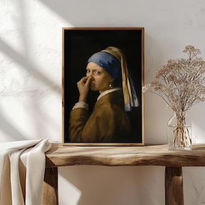 May include: A framed wall art print featuring a portrait of a person wearing a blue headscarf and a brown jacket, holding their nose with one hand. The person has a pearl earring in their visible ear and is set against a dark, solid background.
