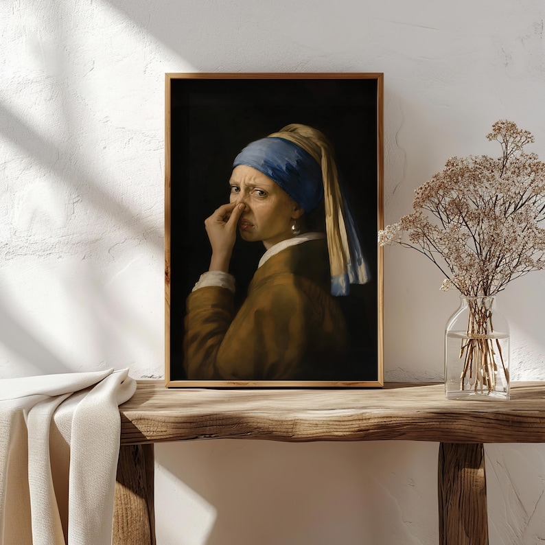 May include: A framed wall art print featuring a portrait of a person wearing a blue headscarf and a brown jacket, holding their nose with one hand. The person has a pearl earring in their visible ear and is set against a dark, solid background.
