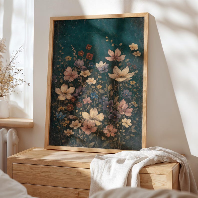 May include: A framed floral print featuring a variety of flowers in shades of cream, pink, purple, and yellow against a dark teal background. The artwork is displayed on a wooden dresser, with a white blanket draped over it.