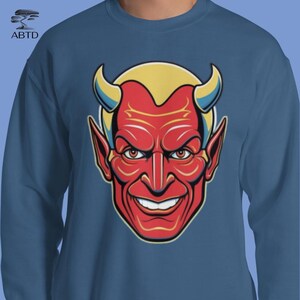 Puede incluir: Sudadera azul con un gráfico de cara de diablo rojo. El diablo tiene pelo amarillo, cuernos azules y una amplia sonrisa. El gráfico está centrado en el pecho de la sudadera, que tiene cuello redondo.
