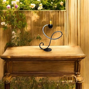 May include: A black metal candle holder with a curved design. The holder has a gold-colored candle on top and sits on a wooden table.