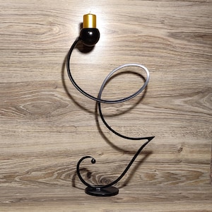 May include: A black metal candle holder with a curved design. The holder has a single candle cup and a round base. A gold candle is in the candle cup.
