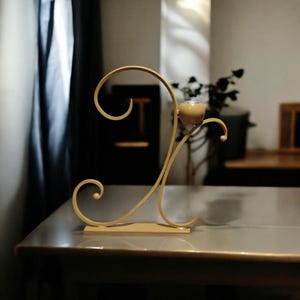 May include: A gold metal candle holder with a decorative, swirling design. The holder has a round top and a base with a flat surface.