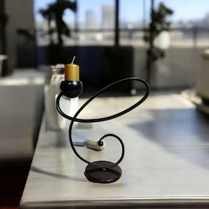 May include: A black metal candle holder with a swirling design. The holder has a round base and a single candle holder on top. A gold candle is in the holder.