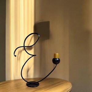 May include: Black metal candle holder shaped like the letter 'E' with a single candle in a black holder. The candle is gold.