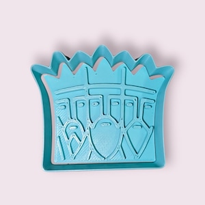 Puede incluir: Cortador de galletas de plástico azul claro con la forma de tres reyes con coronas. El cortador tiene un diseño detallado de las caras y barbas de los reyes. Sobre un fondo rosa claro.