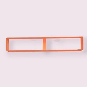 Puede incluir: Un cortador de galletas rectangular de color naranja con un marco de metal fino. El diseño presenta una forma horizontal larga y estrecha dividida en dos secciones iguales por una barra vertical en el centro, creando dos aberturas idénticas.