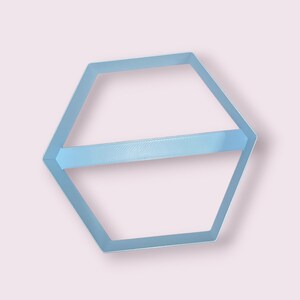 Puede incluir: Un estante hexagonal de color azul claro con una sola barra horizontal que atraviesa el centro. La estructura está hecha de un material sólido y liso con un marco geométrico limpio que crea dos secciones triangulares abiertas en el hexágono.