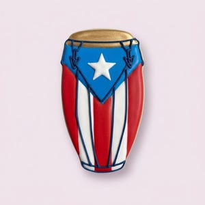 May include: A conga drum-shaped cookie decorated with the Puerto Rican flag. The cookie features a gold top, a blue triangle with a white star, red stripes, and white stripes. The edges are outlined in blue.