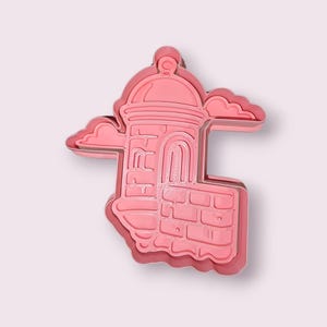 May include: A pink cookie cutter shaped like a building with a domed roof, arched doorway, and brickwork. Two cloud shapes are on either side. The cutter has a layered design.