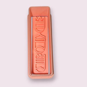 Puede incluir: Un cortador de galletas rectangular de color cobre. El cortador tiene un diseño en relieve con la palabra "MIDTOWN" en orientación vertical. El cortador es de metal y tiene un diseño sencillo y limpio.