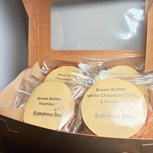 May include: A brown cardboard box with a clear window, filled with individually wrapped cookies. The cookies have round labels that read "Brown Butter Tiramisu" and "Brown Butter White Chocolate Chip & Guava."