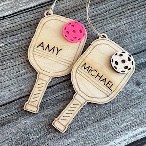 May include: Two wooden pickleball paddle ornaments, one with "AMY" and a pink ball, the other with "MICHAEL" and a black and white ball. Both have a string for hanging.