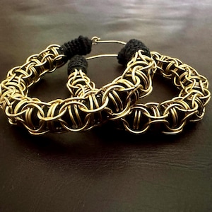 May include: Two gold-colored hoop earrings with a woven chainmail design. The earrings have a black fabric accent at the top. The earrings are displayed on a dark brown surface.