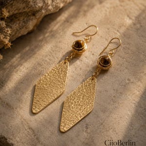 Può includere: Un paio di orecchini pendenti color oro con un ciondolo a forma di diamante testurizzato sospeso da una perla occhio di tigre marrone avvolta in filo metallico. Gli orecchini includono un gancio e il testo CiioBerlin jewelry.