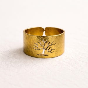 Könnte beinhalten: Ein breiter, goldfarbener Ring mit gehämmertem Muster. Der Ring hat ein ausgeschnittenes Design eines Baumes. Der Ring ist hinten offen, was eine Größenanpassung ermöglicht. Der Ring ist vor einem weißen Hintergrund platziert.