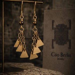 Può includere: Un paio di orecchini pendenti color oro con una perla occhio di tigre marrone, delicate catenelle dorate e tre piccoli ciondoli triangolari testurizzati. Accanto agli orecchini c'è un cartoncino con il testo Ciio Berlin Jewelry.