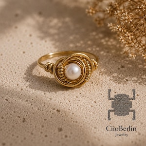Könnte beinhalten: Ein goldfarbener, drahtgewickelter Ring mit einer zentralen weißen Perle. Der Metalldraht ist in einem kreisförmigen Muster um die Perle gewickelt, was ein strukturiertes, geschichtetes Design erzeugt. Das Logo von CiioBerlin Jewelry ist unten rechts.