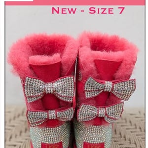 May include: Customized UGG Meilani boots in size 7, featuring a vibrant red exterior with sparkling rhinestone bows and accents. The boots have a plush, pink interior and the UGG logo on the heel.