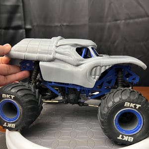 Op de afbeelding: Een grijs 3D-geprint monstertruckmodel met blauwe wielen en "BKT" gedrukt op de banden. De truck heeft een gedetailleerd carrosserie met een gebeeldhouwd hoofd en een dak.