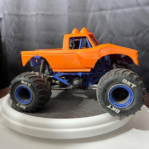 Op de afbeelding: Een oranje monstertruck speelgoed met blauwe accenten. De truck heeft grote, zwarte banden met "BKT" branding en blauwe velgen. De carrosserie is oranje en het chassis is blauw. Het speelgoed staat op een wit platform.