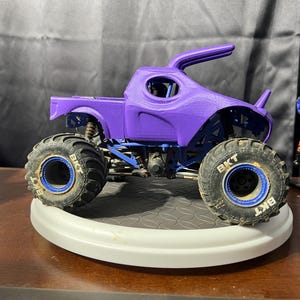 Op de afbeelding: Een paars monstertruckspeelgoed met grote, robuuste banden en blauwe accenten. De truck heeft een uniek cabinedesign en wordt tentoongesteld op een wit platform. De banden hebben de tekst "BKT".
