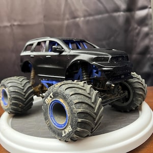Op de afbeelding: Een zwart monstertruckspeelgoed met grote, getextureerde banden en blauwe accenten. De banden hebben de tekst "BKT". De truck staat op een witte draaiende display.