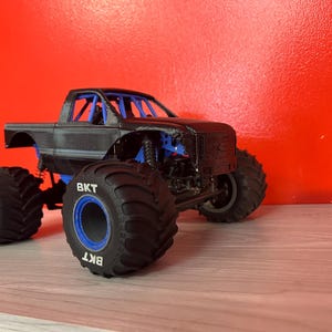 Op de afbeelding: Zwarte monstertruck met blauwe wielen en "BKT" gedrukt op de banden. De truck heeft een zwart carrosserie en een rolkooi.
