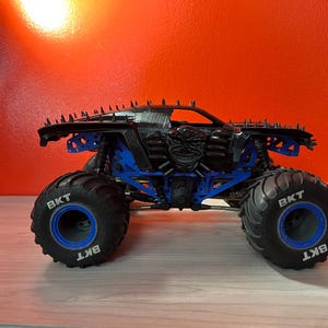 Op de afbeelding: Zwarte monstertruck met blauwe wielen en een schedelprint aan de zijkant. De wielen zijn gelabeld met "BKT".