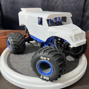 Op de afbeelding: Witte, ongeverfde, plastic monstertruck met blauwe wielen en "BKT" gedrukt op de banden. De truck heeft een cabine met een voorruit en een rolkooi.