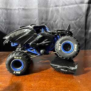 Op de afbeelding: Een zwart en blauw monstertruck speelgoed met grote banden. De truck heeft een gedetailleerd ontwerp met een puntige voorkant en een blauw chassis. De banden hebben de letters "BKT" erop. Het speelgoed staat op een houten oppervlak.