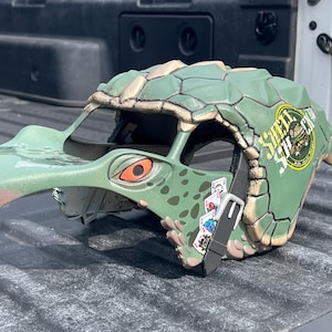 May include: A green and tan bird-head mask with an orange eye and a long beak. The mask has a textured surface and the text "SMALL SHACK" on the side. A playing card design is visible on the side.