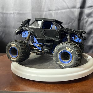 Op de afbeelding: Een zwart en blauw monstertruck speelgoed met grote, robuuste banden. De banden hebben de tekst "BKT" erop. De truck staat op een wit, cirkelvormig platform.