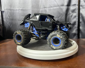 Puede incluir: Un juguete de monster truck negro y azul con neumáticos grandes y resistentes. Los neumáticos tienen el texto "BKT". El camión está sobre una plataforma blanca y circular.