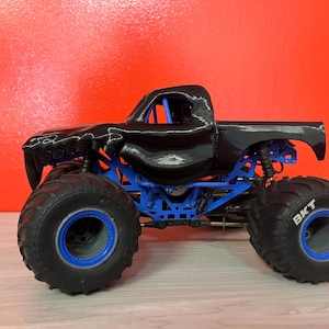 Op de afbeelding: Een zwart monstertruckspeelgoed met blauwe ophanging en grote zwarte banden. De banden hebben de tekst "BKT" in het wit. De truck staat tegen een rode achtergrond.