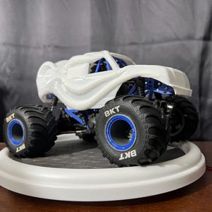 Op de afbeelding: Een witte monstertruck met blauwe wielen en "BKT" gedrukt op de banden. De truck staat op een wit rond platform.