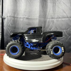 Op de afbeelding: Zwart monstertruck speelgoed met blauwe wielen en "BKT" gedrukt op de banden. De truck heeft een zwart-witte verfbeurt en een blauwe rolkooi.