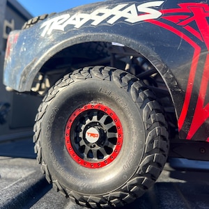 May include: Close-up of a black and red off-road vehicle tire with a red rim and the TRD logo. The tire is mounted on a black wheel. The vehicle's body is black with red graphics and the word "TRAXXAS" in white.