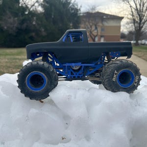 Op de afbeelding: Een zwarte monstertruck met blauwe accenten en oversized banden. De truck staat op een met sneeuw bedekt oppervlak. De banden hebben de tekst "BKT" op de zijkanten.