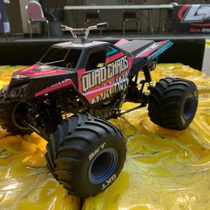Op de afbeelding: Een zwarte en roze monstertruck met de woorden "QUAD CHAOS" erop gedrukt. De truck heeft grote zwarte banden met blauwe velgen en staat op een gele ondergrond.