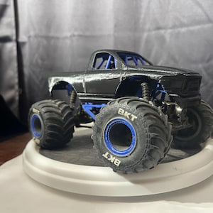 May include: A black monster truck toy with large, textured tires and blue accents. The truck has a detailed body and is displayed on a white platform. The tires have the text "BKT" on them.