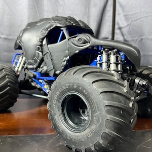 May include: A black and blue monster truck toy with large Goodyear tires. The truck features a detailed, stylized eagle head design on the front and a blue roll cage. The tires have a tread pattern for off-road use.