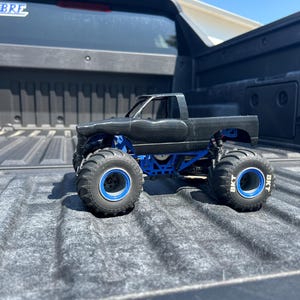 May include: A black monster truck toy with blue accents and large tires. The truck is sitting in the bed of a pickup truck. The tires have the text "BKT" on them.
