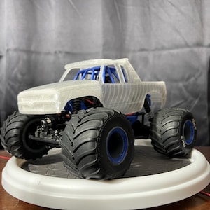 Op de afbeelding: Een wit 3D-geprint monstertruckmodel met blauwe wielen. De truck heeft een rolkooi en een laadbak.