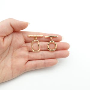 Op de afbeelding: Twee goudkleurige tuimelsluitingen, een rechthoekige en een cirkelvormige, worden getoond. Elke sluiting heeft een horizontale balk die aan een decoratieve lus is bevestigd. De sluitingen worden tegen een witte achtergrond getoond.